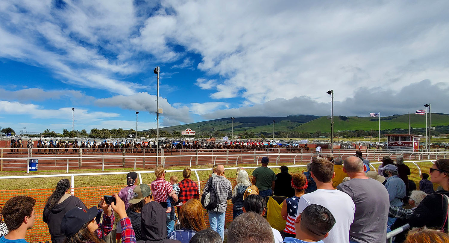 A wide view of the Parker Ranch Rodeo Arena in Waimea, showing a crowd of spectators watching a long lineup of paniolo on horseback.